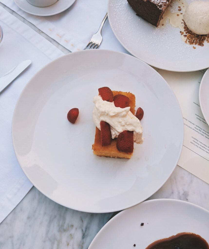 Picture of a slice of olive oil cake with whipped cream and strawberries. It is on a white plate.
